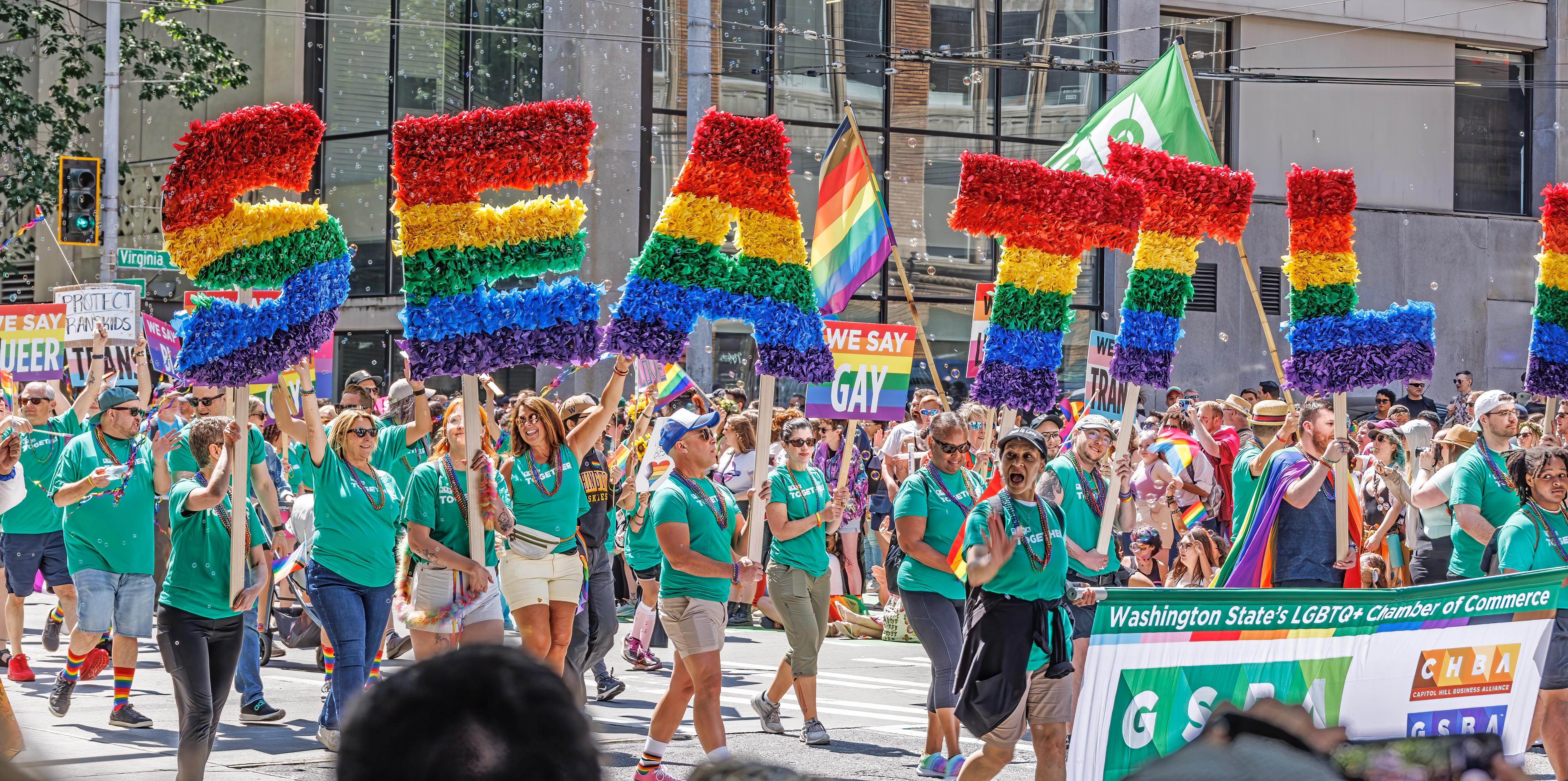 Photo of a pride parade at Mirabella Seattle. Participants are wearing teal t-shirts and holding up rainbow decorative letters that read Seattle.