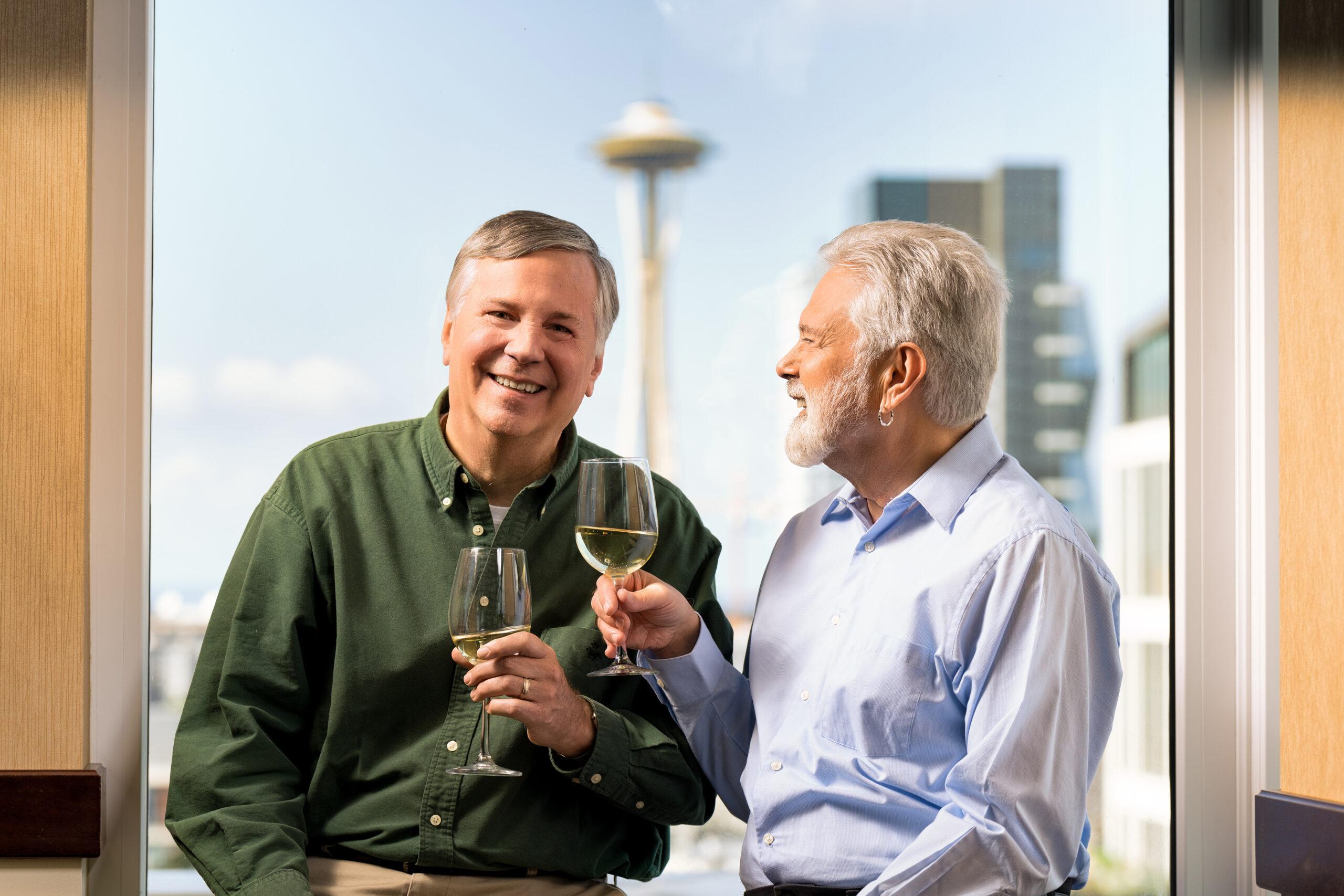 Two men smiling and sipping wine, sitting in front of a window with a view of the Space Needle.