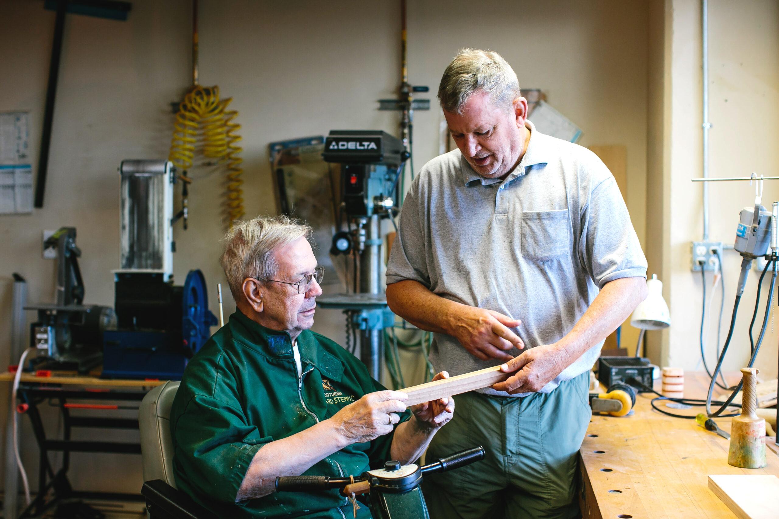 Two older men examine a piece of wood in a wood shop.