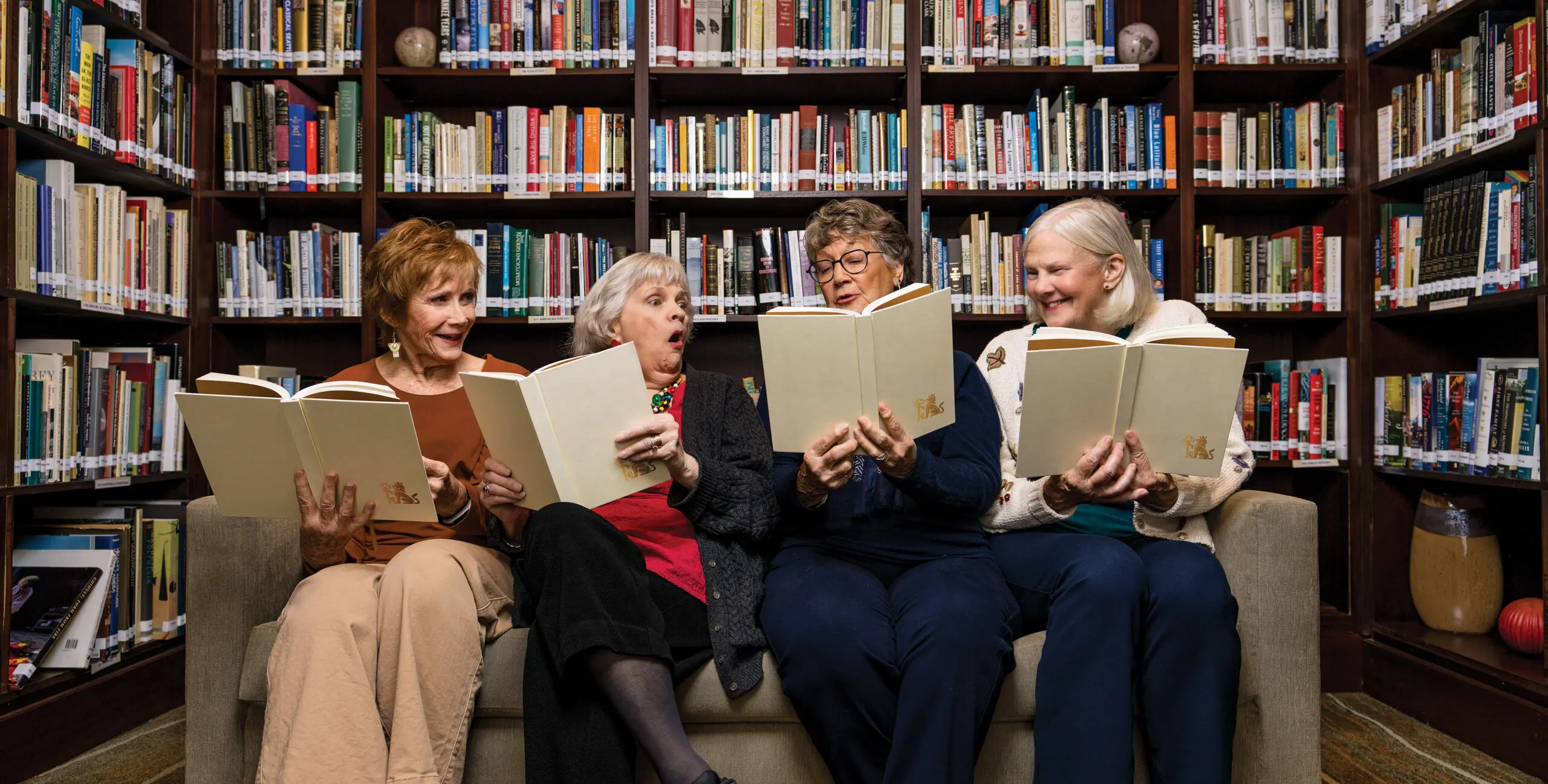 Four women sit on a couch holding books with humorous and fun expressions. They are sitting in front of a large bookshelf.
