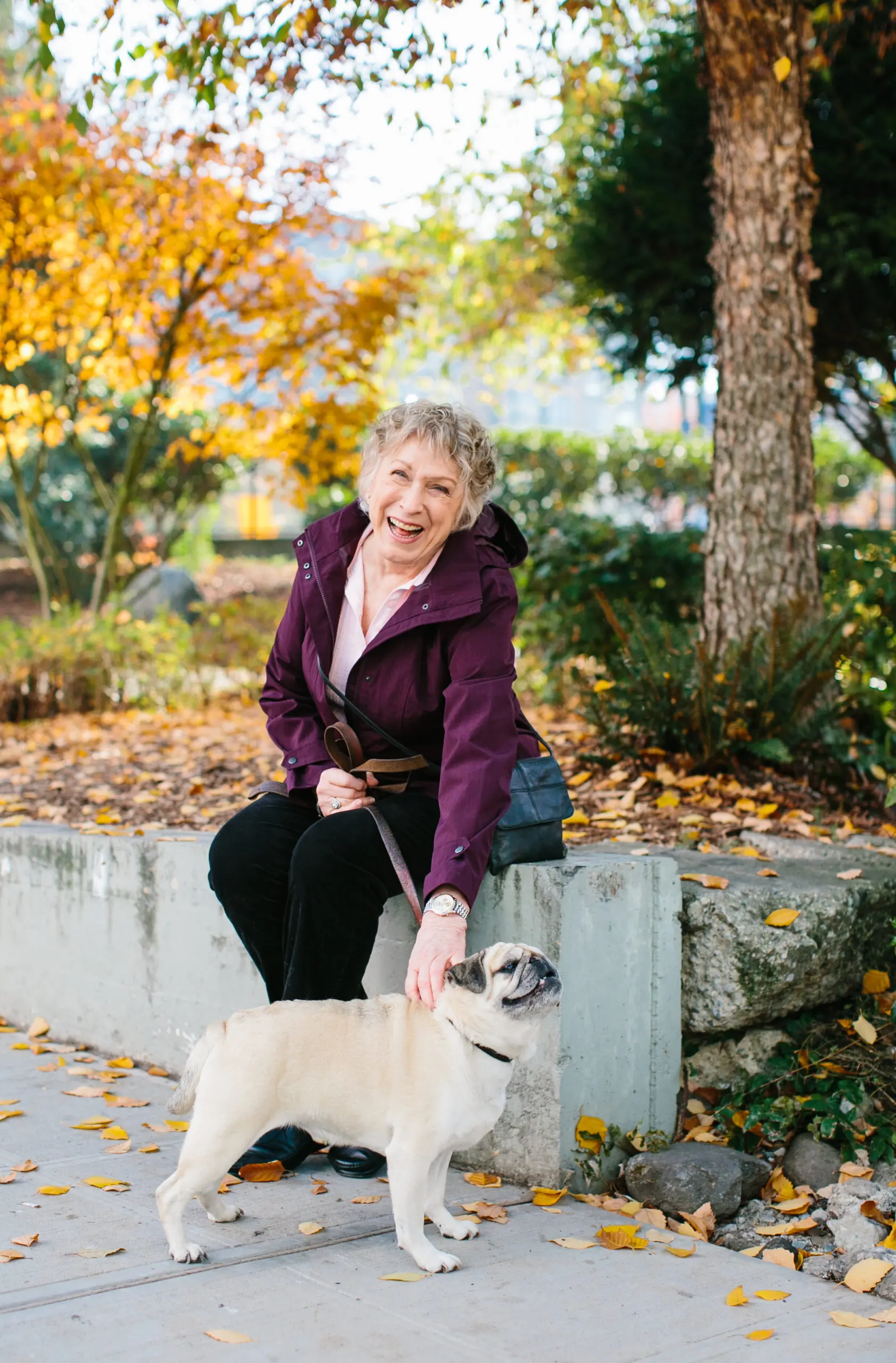 A happy woman sits outdoors and pets a dog, with beautiful fall colors in the background.