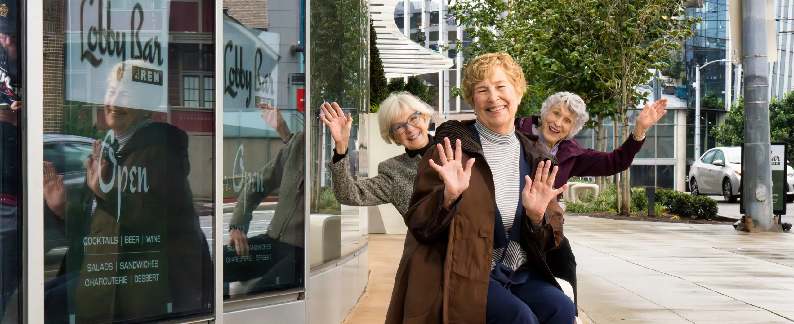 Three smiling women doing a fun pose for a photo outdoors. Their hands are outstretched.